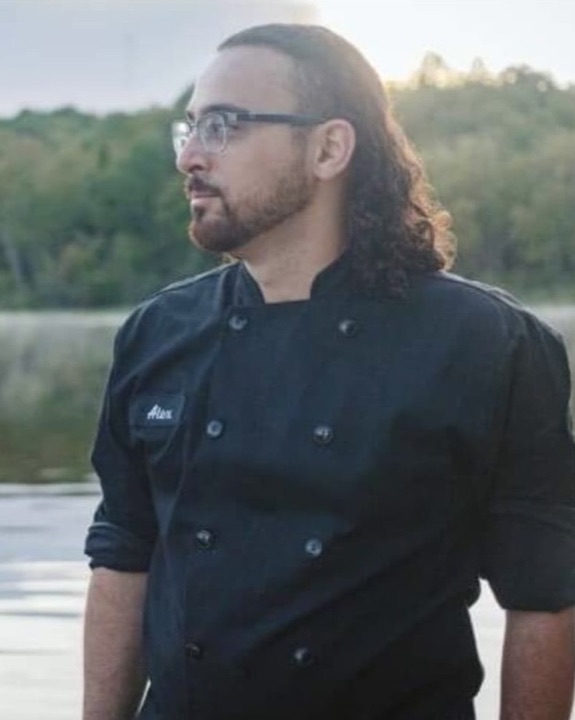Alex, in a black chef's coat, by a lake at dusk